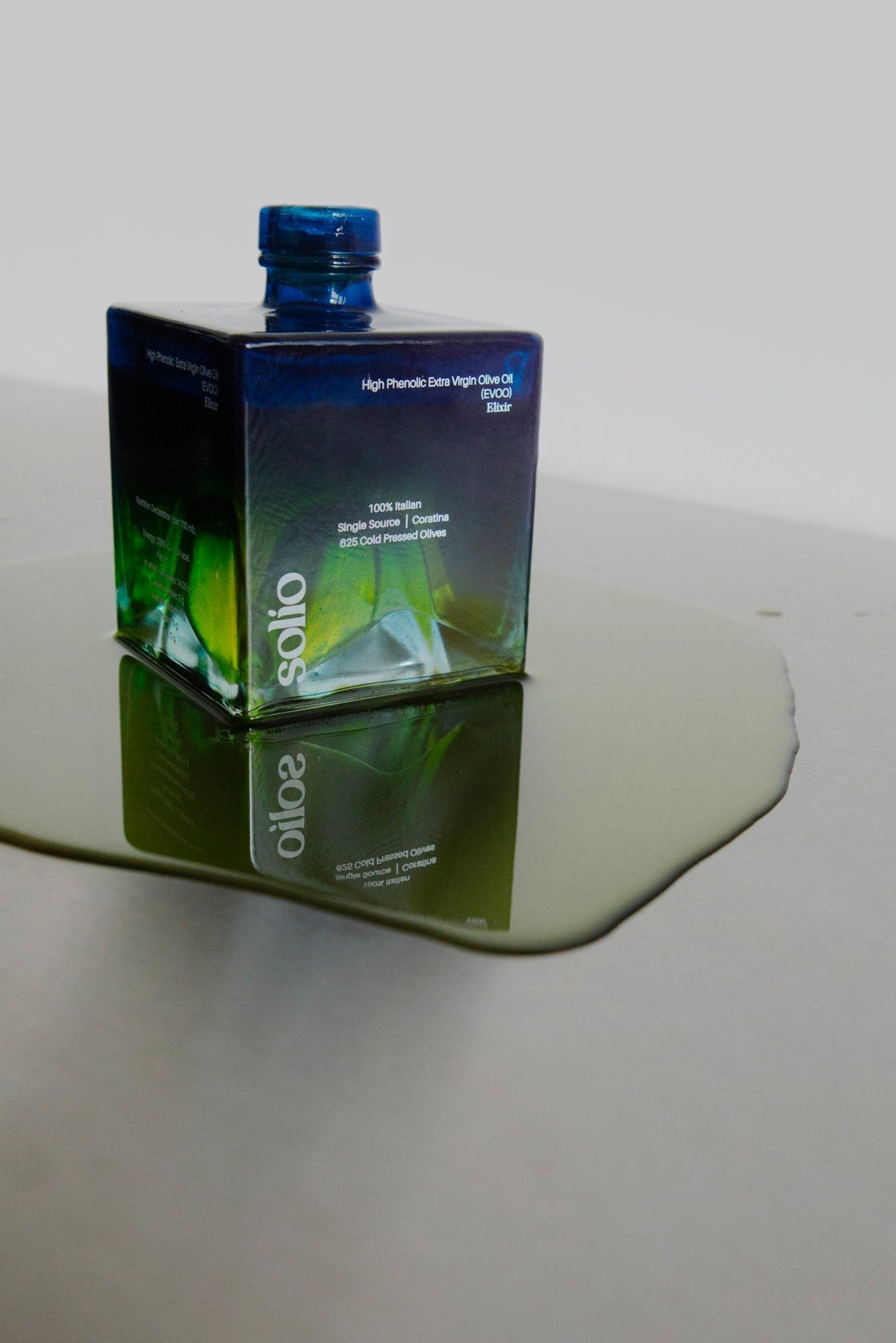 Würfelförmige Solio Flasche steht in einer Pfütze aus grünem Olivenöl auf Edelstahl
